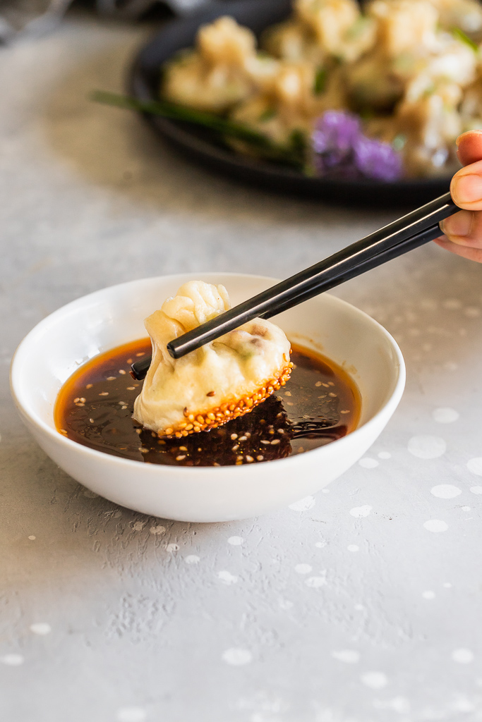 a vegan dumpling being dipped into a white bowl of teriyaki sauce with chopsticks
