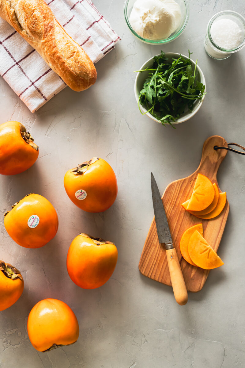 a french baguette on a tea towel, whole persimmons, sliced persimmons on a cutting board with a knife, a bowl of arugula, and glass jars filled with flaky salt and burrata cheese