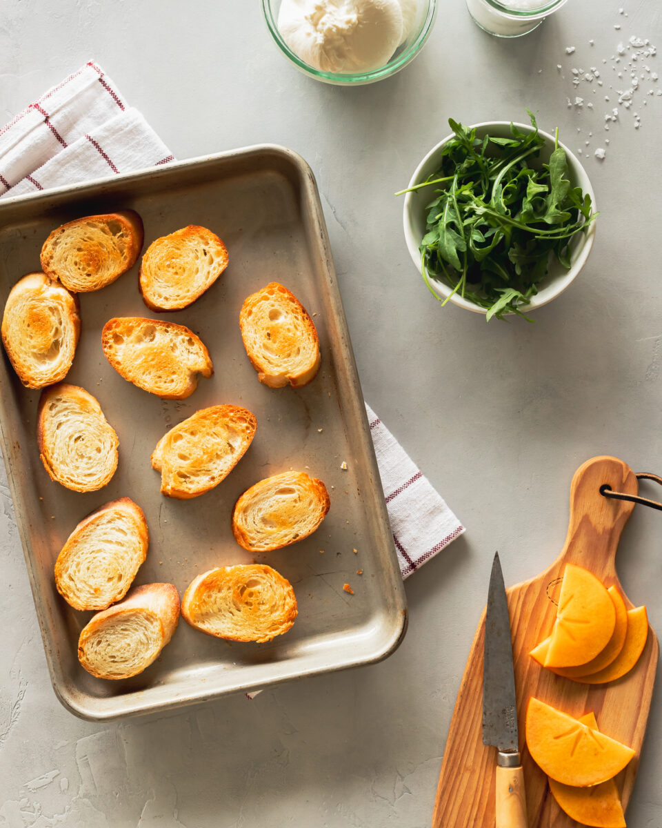 a baking tray of crostini, a bowl of arugula, a jar of burrata and a wooden cutting board with sliced persimmons on a grey surface