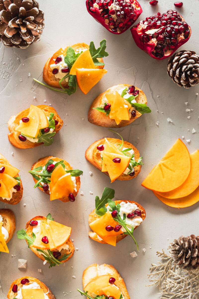 persimmon bruschetta on a grey surface with pine cones bordering the image