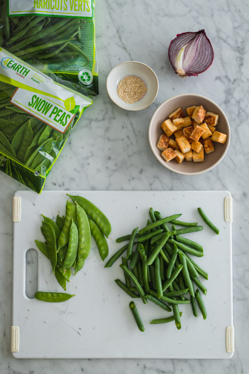 a marble countertop with a green beans and snow peas on cutting board next to a bowl of cooked tofu, half a red onion and a bowl of sesame seeds