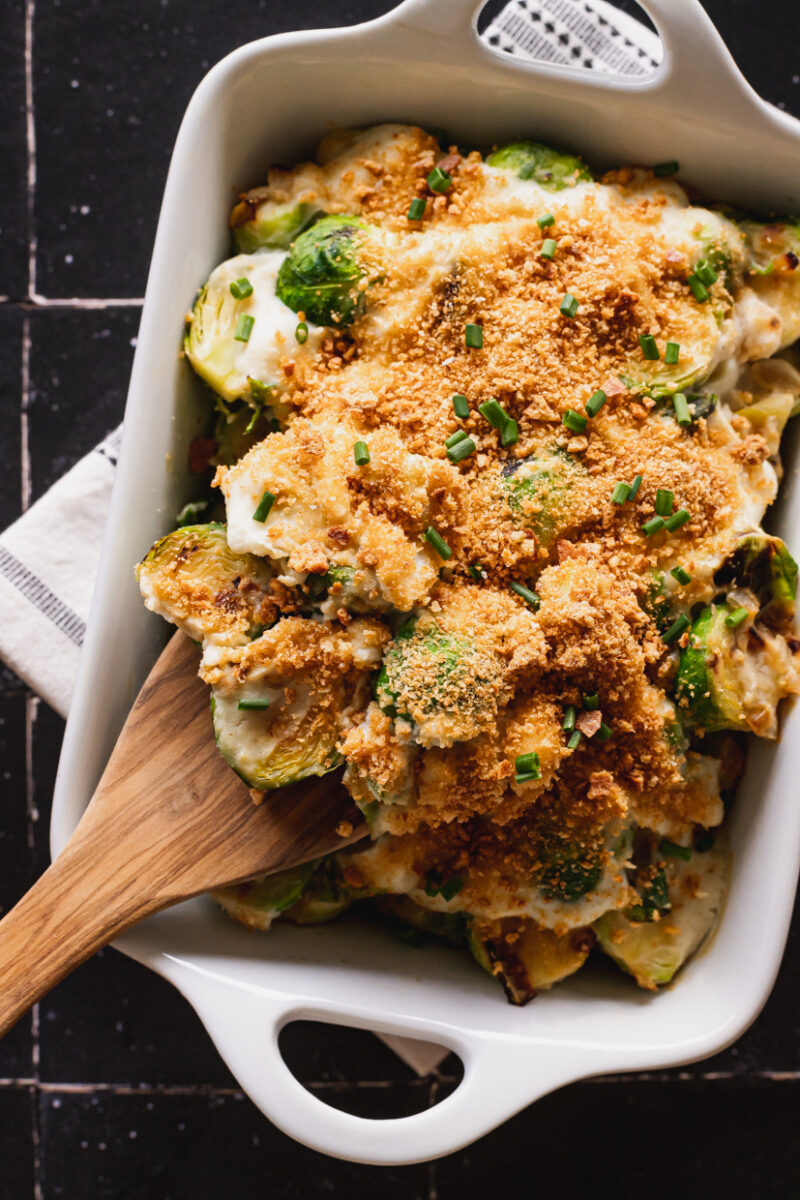 Brussels Sprouts Gratin in a white casserole dish and a wooden spoon scooping out a portion