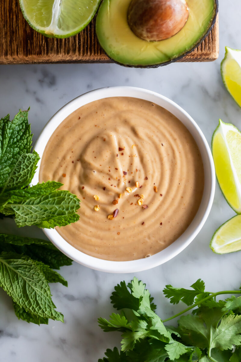 overhead view of a bowl of peanut dipping sauce on a marble table surrounded by bunches of fresh mint and cilantro, lime slices and an avocado half