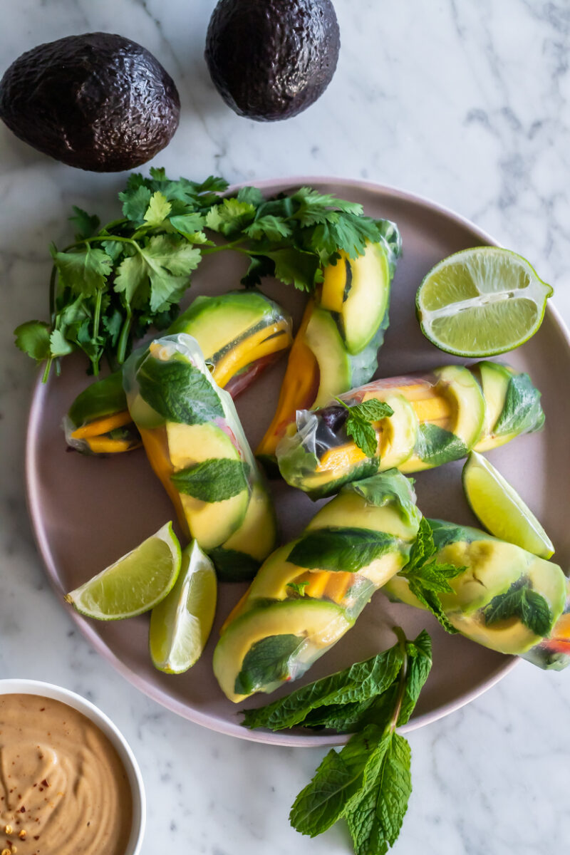 aerial view of avocado summer rolls with mango, mint and lime on a light pink ceramic platter with whole avocados next to it